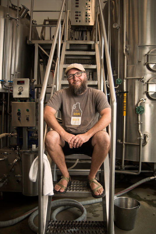 A bearded man wearing a cap, sits on a metal staircase. He is in a brewery.