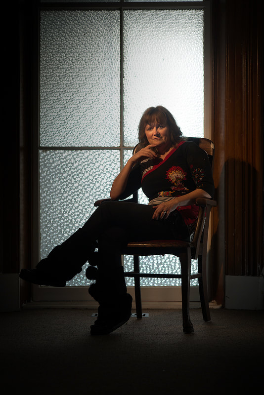A woman is sitting on a chair in a darkened room. She sits in front of a textured window.