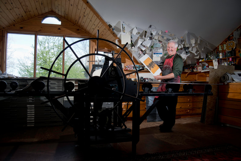 A mature man stands in an art studio, holding an art print he has made. He leans against a large printing press.