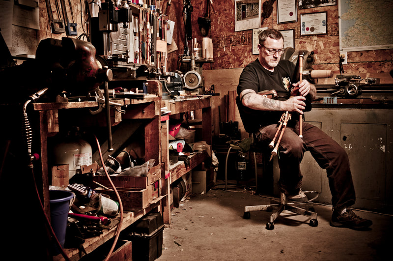 A bagpipe player sits in a workshop, playing the pipes. Shot by Peterborough professional photographer, Mike Taylor.