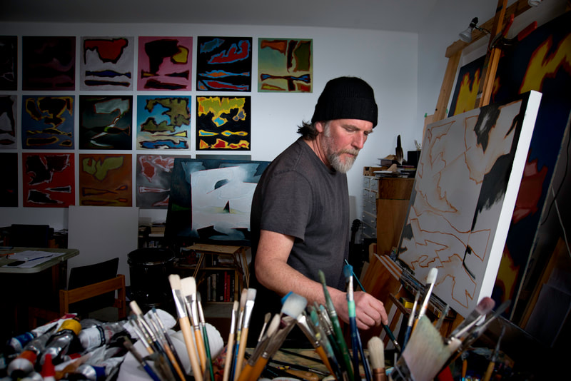 A man with a black cap paints a picture in his art studio.