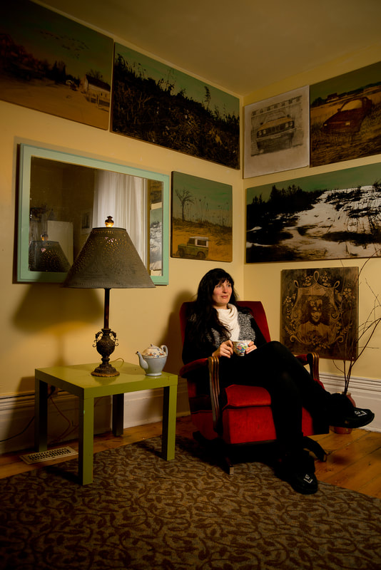 A woman sits on a red chair in the corner of a yellow walled room. Many painting cover the walls.