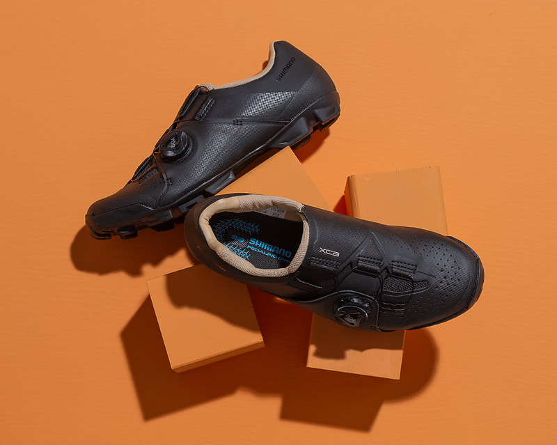 A pair of black and tan cycling shoes are displayed on an orange surface. Shot in studio by photographer Mike Taylor.