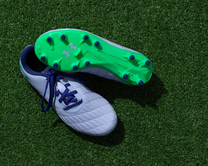 A pair of soccer cleats in the colours of blue and green, rest on a brightly lit green grass surface. Photographed by professional photographer Mike Taylor.